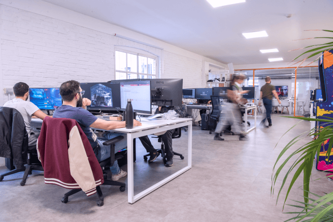 An open office space with team members working at desks with multiple monitors, and others walking in the background.