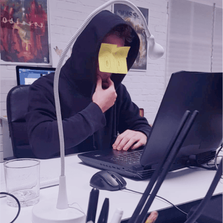 A person at a desk with a laptop, wearing a hoodie and holding a Post-it note with a drawing over their face.