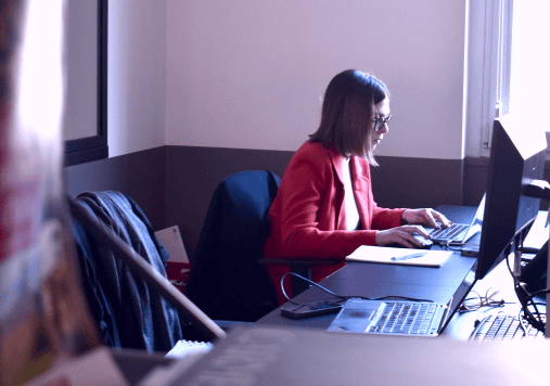 A team member working at a desk in an office environment, focused on a computer screen, wearing a red blazer.