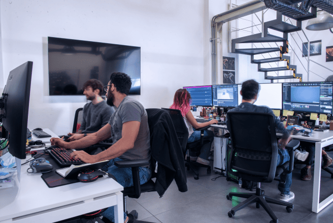 A group of team members working at desks in a modern office, with a staircase and a large screen in the background.