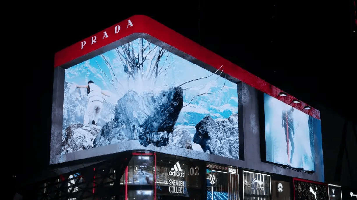 Close-up di un cartellone pubblicitario 3D di Prada Linea Rossa raffigurante un paesaggio montano dinamico, visto di notte.