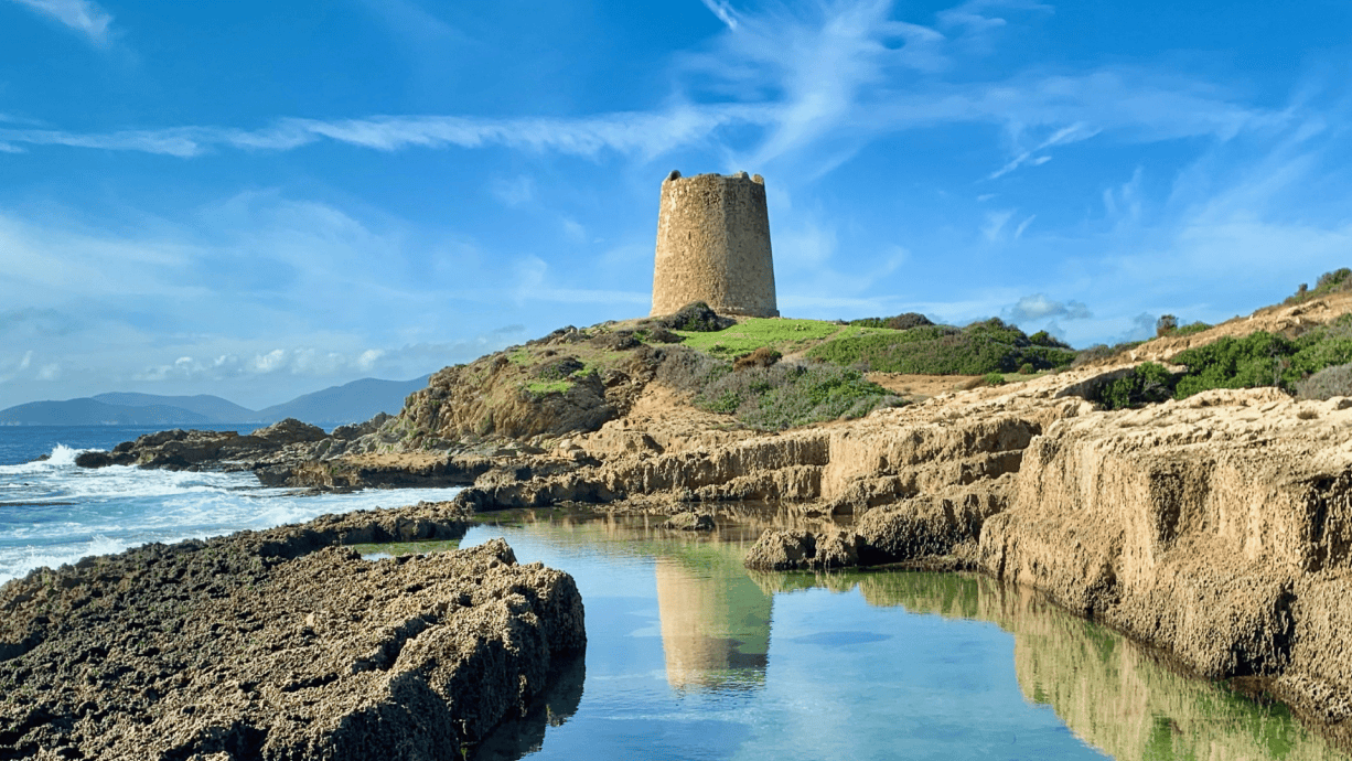 Stone watchtower on a rocky Sardinian coastline, surrounded by clear blue skies and ocean waves.